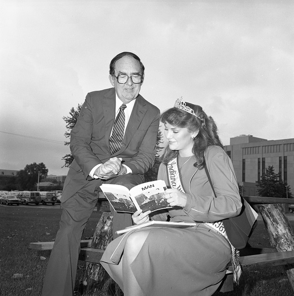 Young woman with a tiara and a sash reading, 'Indiana,' holds open a book for an older man in a suit who is seated above her