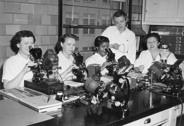 Four women looking at the camera with microscopes in front of them. Another woman in a lab coat stands over them