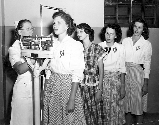 Four young women line up before a mechanical scale with a nurse taking one's height