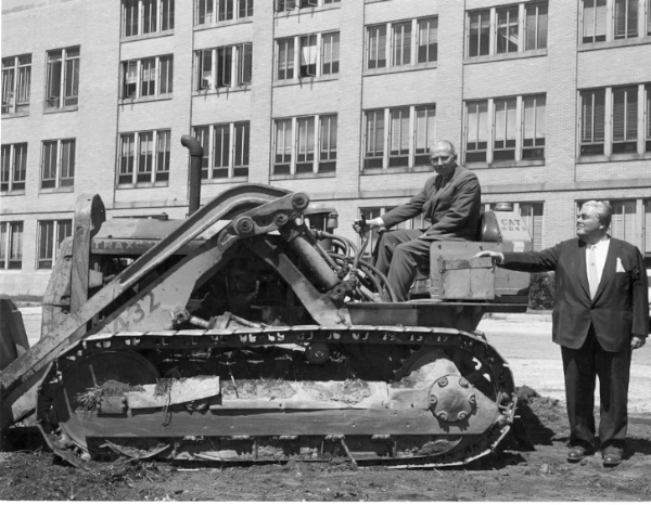 Black and white photograph. A multi-storied college campus building extends up off the frame in the background. The building has many windows. There is the side shot of a large bulldozer in the middle of the picture. An elderly white man wearing a suit is sitting on the bulldozer in the driver's seat facing the photographer. A large white man in a suit stands to the right side of the bulldozer looking at the other man. His arm stretches out and rests on the bulldozer.