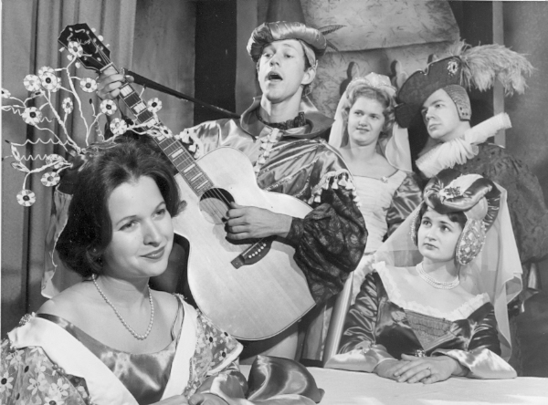 A woman with an ornate headdress looks to the side while a man plays guitar. Three other people stare at the man. They all wear shakespearian style clothing
