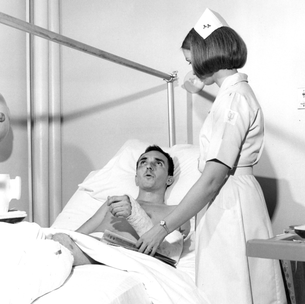 Man laying in a hospital bed with an arm cast looking up at the ceiling while a nurse watches him