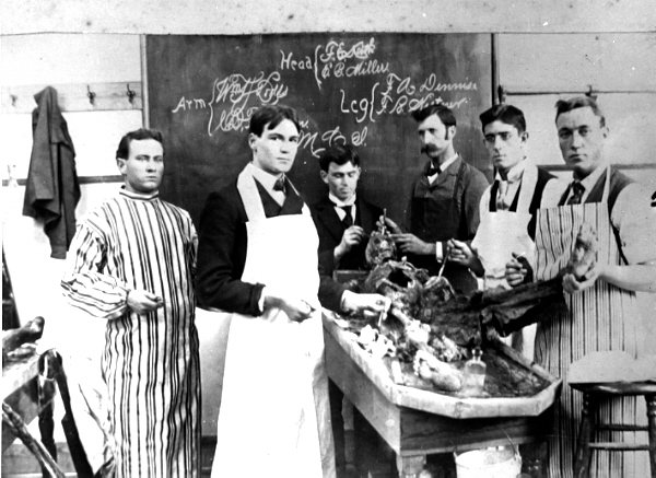 Black and white photograph. A medical school classroom from the early 20th century. Six young white men with serious expressions  stand in front of a chalkboard with notes drawn on it. One of the men has a handle bar mustache. They are surrounding a lab table with cadaver parts laid out on top. The men are wearing clothing consistent with the era, and they are wearing lab coats over the top. The man on the right is holding up a severed leg. 