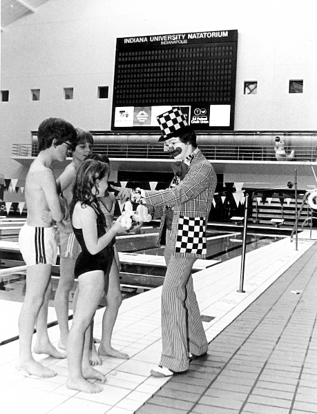 A man in clown makeup is point at the nose of a young girl with two young boys watching. They are standing next to an indoor swimming pool and the children are in swim clothes