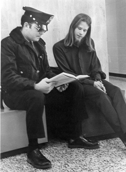 Man with long hair and goatee is seated next to a cop in sunglasses, looking at a book together