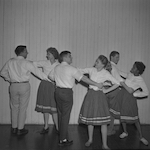 Black and white photograph. Three couples are dancing together, with the men on the left of their partners and the women on the right. The women have a hand on their partner's shoulder and the other in a fist on their hips. The men are posed similarly, but with their hand on their partner's waist. Everyone is wearing a button up shirt, with the women wearing skirts with two horizontal white lines near the bottom. On the right hand side of the picture a smiling woman is leaning on a table watching them dance. On the table is a sash.